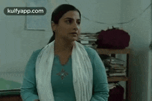 a woman in a blue shirt and white scarf is standing in a room with a stack of books on a shelf .