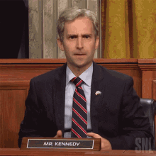 a man in a suit and tie is sitting at a desk with a name plate that says mr. kennedy