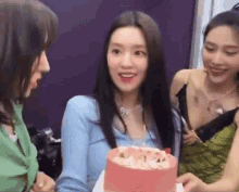 a woman in a blue shirt is holding a pink cake while two other women look on .