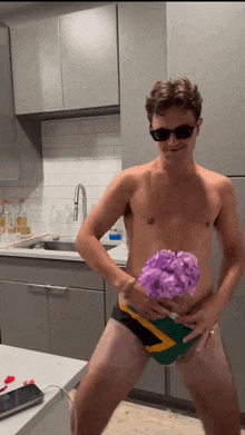 a shirtless man holding a vase of purple flowers in a kitchen