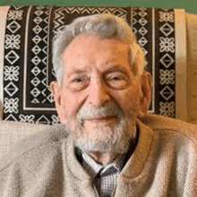 an older man with a beard is sitting in a chair and smiling .