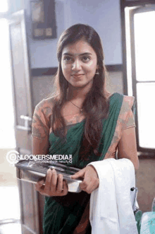 a woman in a green and orange saree is holding a book and a white coat .