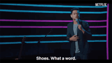 a man in a suit stands on a stage holding a microphone and says shoes what a word