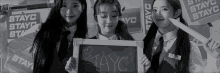 three girls in school uniforms holding a sign that says stayc