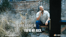 a man sitting on a bench talking on a cell phone with the words " remember you 're my rock " above him