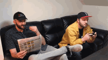 two men are sitting on a couch reading classifieds and one is smoking a cigar