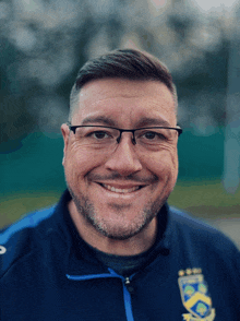 a man wearing glasses and a blue jacket with the letters kc on it smiles for the camera