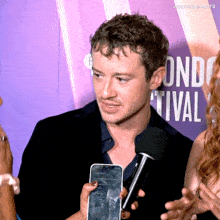 a man talking into a microphone while holding a cell phone in front of a sign that says london festival