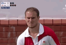 a man in a red and white shirt stands in front of a brick wall with a scoreboard that says scot 8 england 11