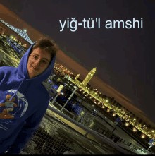 a man wearing a blue hoodie stands in front of a city at night with the words yig-tu'l amshi above him