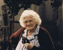 an elderly woman wearing glasses and a red cardigan holds a camera