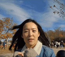 a woman in a denim jacket is standing in a park with her long hair blowing in the wind .