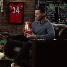 a man sitting on a couch eating a bag of chips in front of a chalkboard that says hall of fame