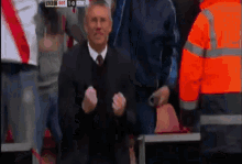 a man in a suit and tie stands in front of a crowd watching a soccer game with the score at 1-0