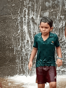 a young boy standing in front of a waterfall wearing a green shirt with a crown on it