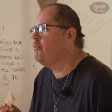 a man wearing glasses and a black shirt is eating a piece of food in front of a white board with writing in spanish