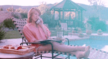 a woman in a pink shirt is laying on a lounge chair by a pool talking on a cell phone