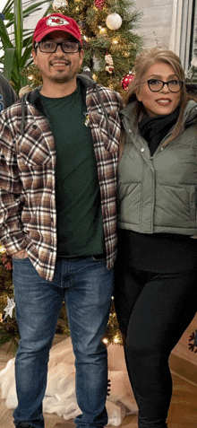 a man wearing a red chiefs hat stands next to a woman wearing glasses in front of a christmas tree