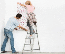 a man and a woman are hanging wallpaper on a wall . the woman is standing on a ladder .