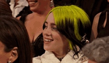 billie eilish is smiling while sitting in a crowd of people with green hair .