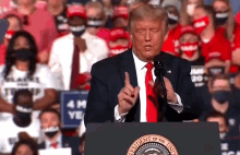 donald trump is giving a speech at a podium in front of a crowd while wearing a red tie .