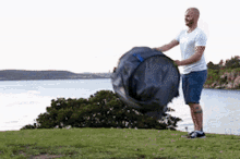 a man in a white shirt is standing on a grassy hill near a body of water holding a large bag