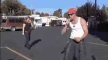 a man in a white tank top and a red bandana is standing in a parking lot with a woman walking behind him .