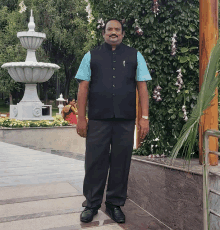 a man in a black vest and blue shirt stands in front of a white fountain