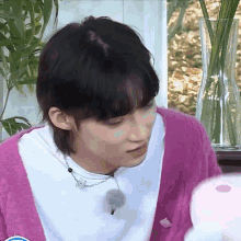 a young man wearing a pink cardigan and a white shirt is sitting in front of a vase of flowers .