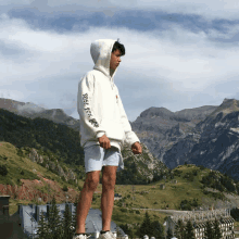 a young man wearing a white hoodie that says raise your head stands in front of a mountain range