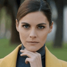 a woman wearing a yellow coat and a black turtleneck has her hand on her chin