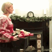 a woman is holding a tray of christmas crackers in front of a fireplace with a clock in the background