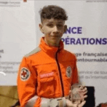 a young man in an orange jacket is holding a trophy in front of a sign .