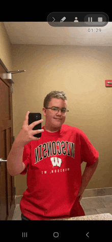 a young man wearing a red wisconsin shirt takes a selfie in a bathroom