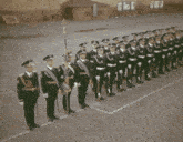 a group of soldiers marching in a line with a flag in front of a building with the letters m and t on the windows