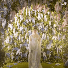 a woman in a white dress stands in front of a wall of flowers with the letters lje below her