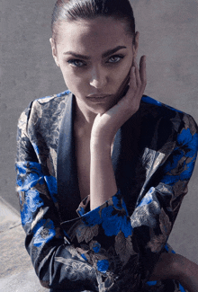 a woman wearing a blue and black floral jacket holds her hand to her chin