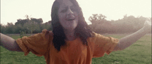 a woman in an orange shirt is standing in a grassy field with her arms outstretched .