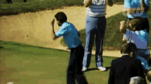 a group of people are standing on a golf course watching a man hit a golf ball out of a bunker .