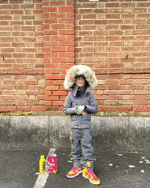 a child wearing a fur hood stands in front of a brick wall next to a bag of bubbles