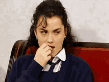 a woman in a blue sweater and tie is sitting on a red couch and covering her nose with her hand .