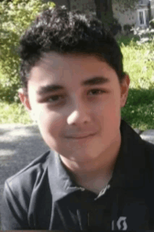 a young boy with curly hair is wearing a black polo shirt and smiling for the camera .