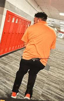 a man in an orange shirt and black pants stands in front of red lockers