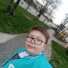 a young boy wearing glasses and a teal jacket is taking a selfie in a park .