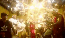 a group of people are standing in a room with balloons and a man wearing an adidas shirt .