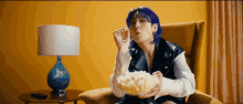 a person with purple hair is sitting in a chair eating popcorn .