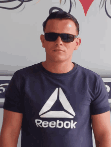 a man wearing sunglasses and a reebok shirt stands in front of a wall