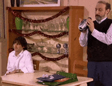 a man is holding a can of soda while a woman sits at a table in front of a shelf full of wine glasses