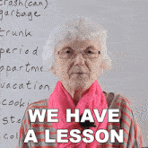 an elderly woman wearing glasses and a pink scarf stands in front of a white board that says we have a lesson