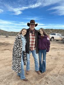 a man in a cowboy hat stands with two girls in a dirt field